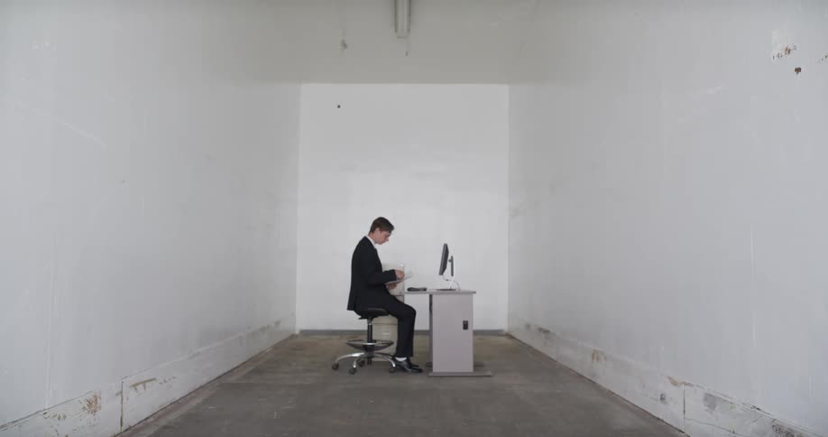 A businessman works at his desk in an empty, sterile white room, reviewing an endless stream of printed paper