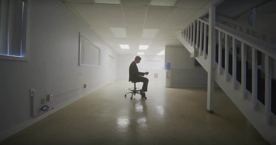 A solitary businessman in a suit works on a laptop while sitting on an office chair in the middle of a vast, empty, white room