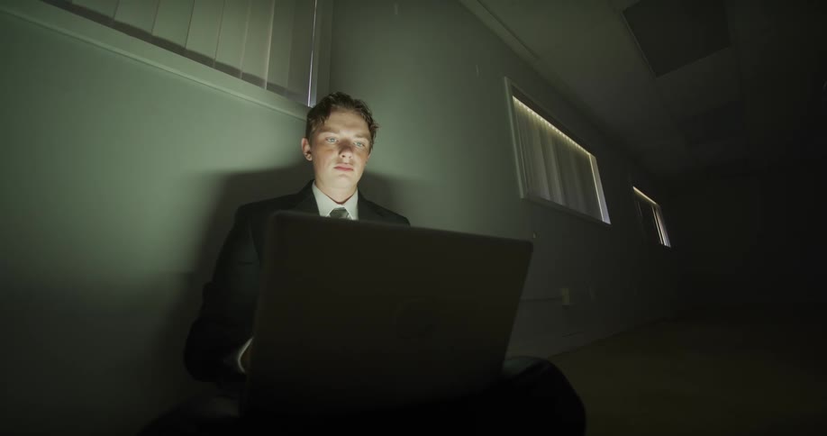 A young man in a business suit sits on the floor of a dark office hallway, intensely focused on his laptop screen
