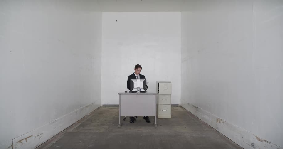 A businessman in a suit works at his desk inside a sterile, empty white room, pulling a file from a cabinet