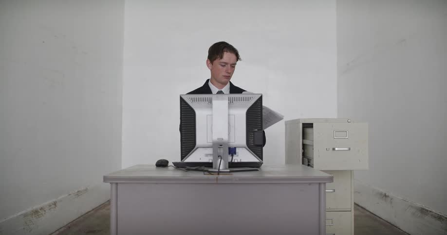 A young businessman retrieves a file from a cabinet, reviews the paperwork at his desk, and files it away in a sterile office environment
