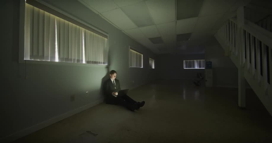 Slow zoom out reveals a solitary businessman working on a laptop, sitting on the floor of a vast, dark, and empty office building