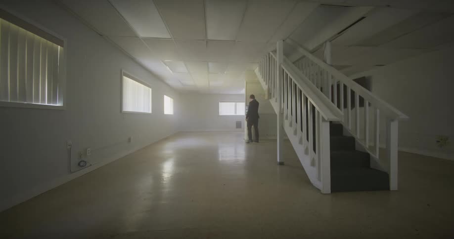 A businessman in a suit walks down the stairs into a large, empty basement and gets a drink of water from a cooler