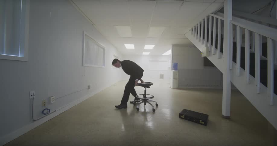 A lone businessman in a suit rights a fallen chair in a vast, empty white room and sits, spinning slowly
