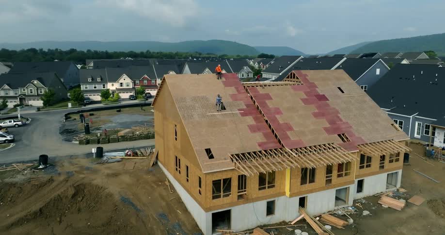 An aerial shot slowly orbits a new townhouse as two construction workers lay plywood sheathing on the roof