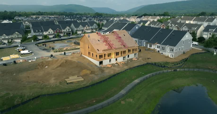Aerial view pulls back from construction workers on the roof of a new home in a suburban development to reveal a scenic pond