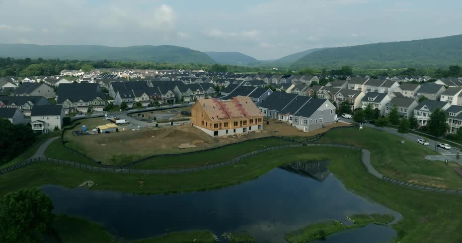 Drone footage slowly flies towards a new home under construction in a modern suburban development with a pond and hills in the background