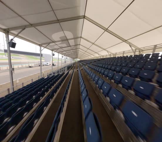 A smooth backward tracking shot through long rows of empty blue seats in a covered grandstand next to a professional racetrack