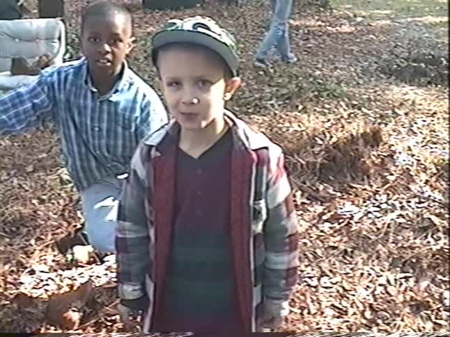 A vintage home video shows two young boys, friends, smiling and talking to the camera while playing outside in a yard covered with autumn leaves