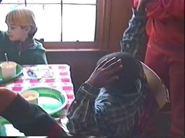 A vintage home movie captures a diverse group of excited children at a birthday party raising their hands for a slice of cake