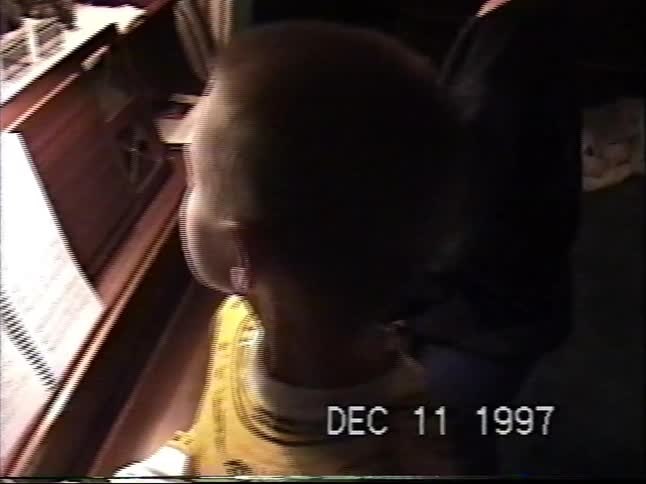 A young boy looks at sheet music at a piano adorned with a festive Christmas village in a 1997 home video