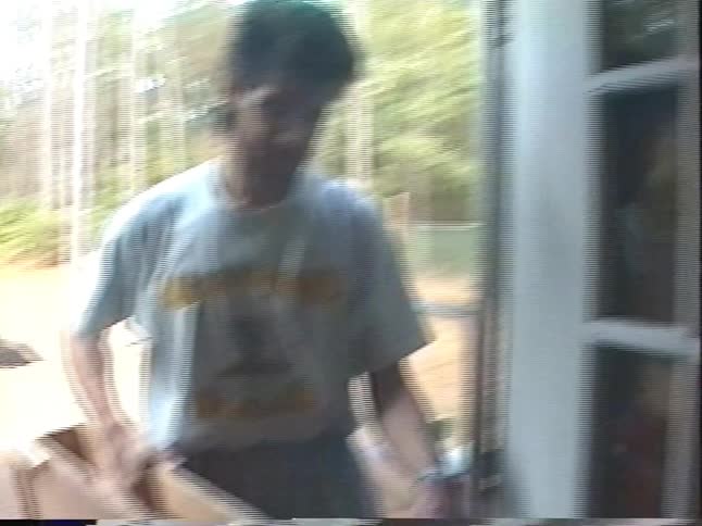 A man with a mullet and beard carries a wooden folding table into his house in a home video from 1997