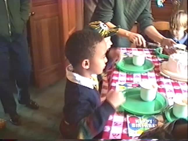 A young boy sits at a table with a checkered tablecloth during a birthday party as an adult cuts the cake in this vintage home video