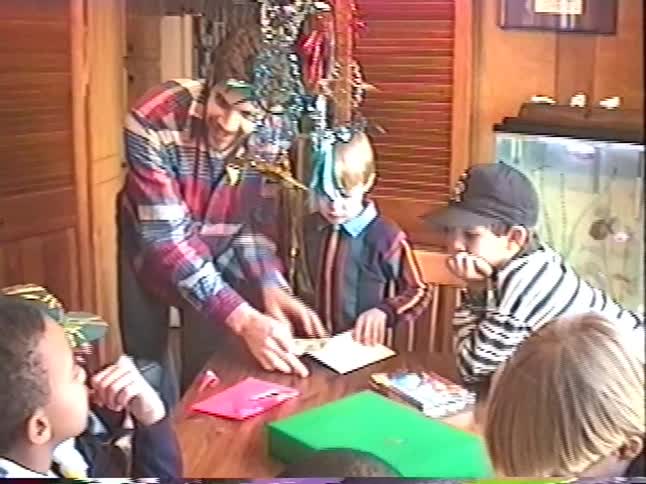 A man with tinsel around his neck performs a magic trick for a group of boys at a festive birthday party in a retro home video