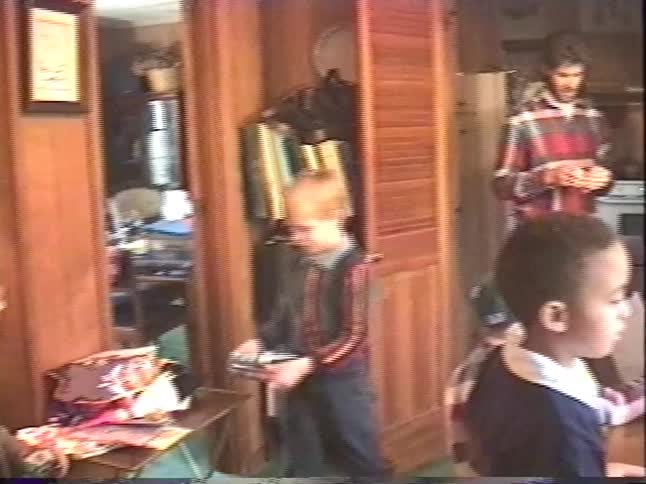 Vintage VHS footage captures a children's party in a cozy home, with kids gathered around a table with new gifts