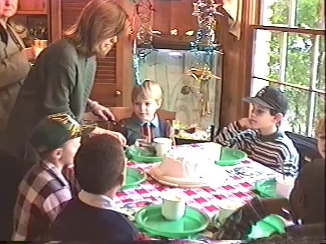 A mother cuts a birthday cake for her son and his friends at a home party as his grandmother captures the moment on camera