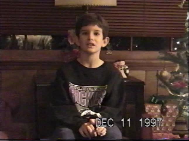 A young boy sits by a brightly lit Christmas tree and speaks to the camera in a nostalgic home video from 1997