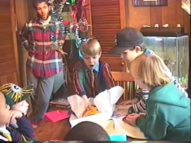 Vintage VHS footage shows a young boy's ecstatic reaction as he opens a gift while his friends and an adult watch at a party
