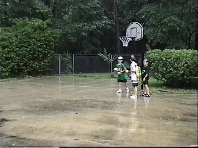 A young boy runs in slow motion and shoots a volleyball into a basketball hoop while playing with friends in the rain in a nostalgic home video