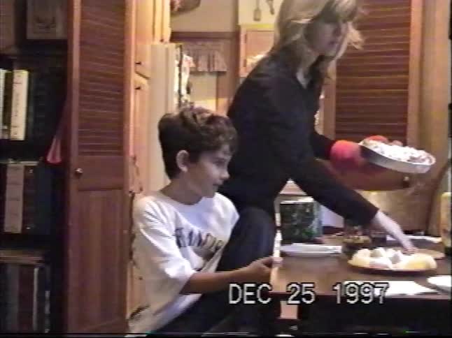 A mother brings a freshly baked dish to the table for her son on Christmas Day in 1997, captured on VHS home video