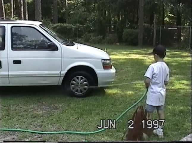 A young boy helps wash the family minivan with a garden hose in the backyard on a summer day in 1997
