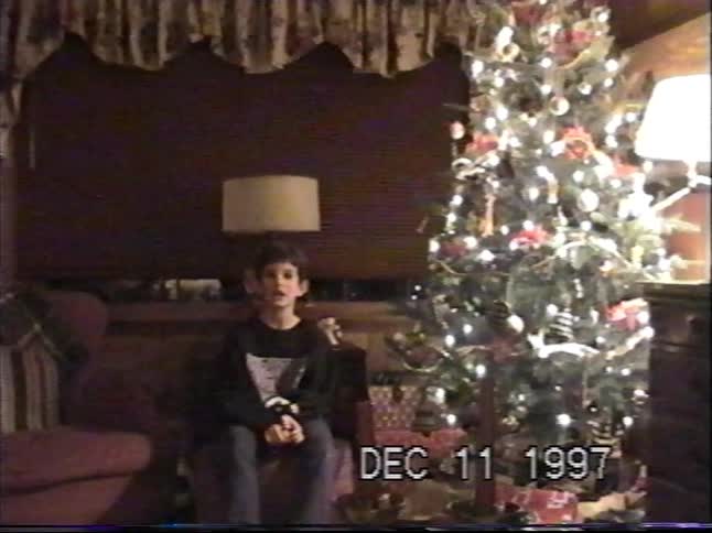 A young boy talks to the camera while sitting next to a brightly lit Christmas tree in this 1997 home video