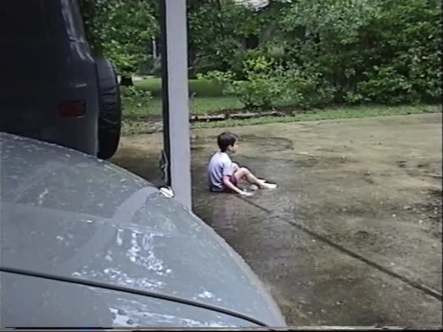 Vintage footage shows a young boy sitting thoughtfully in a rain-soaked driveway as another child runs by