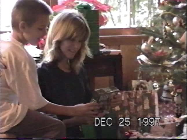 A woman and a young boy open a Christmas present together next to a decorated tree in a 1997 home video