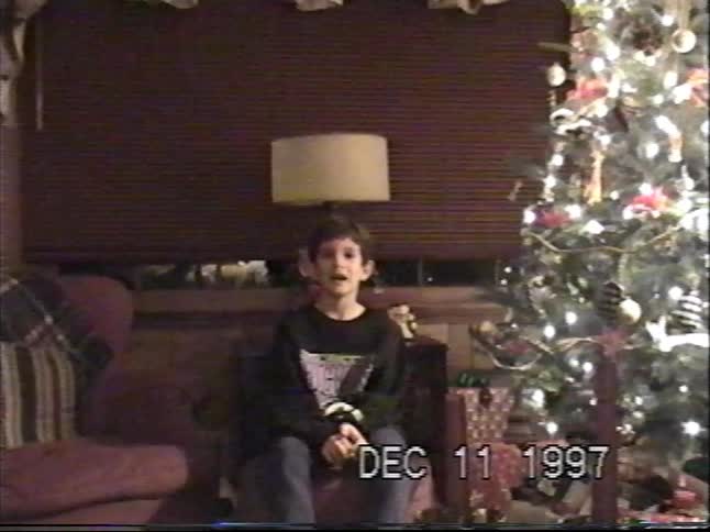 A young boy sits next to a lit Christmas tree and talks to the camera in this vintage home movie from 1997