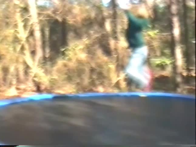Vintage home video captures a young boy with blond hair gleefully jumping and tumbling on a trampoline in a wooded backyard