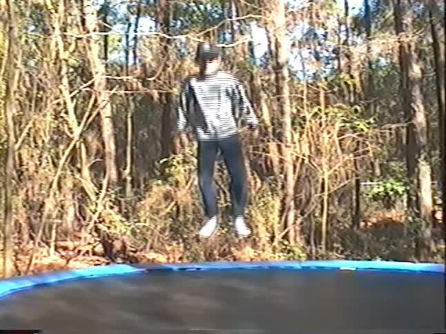 A young boy joyfully jumps on a trampoline in a wooded backyard before the camera pans to show other family members playing catch in this retro home movie clip