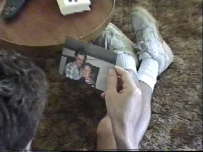 A man sits on a shag carpet and looks at an old photograph of a couple in a vintage home video