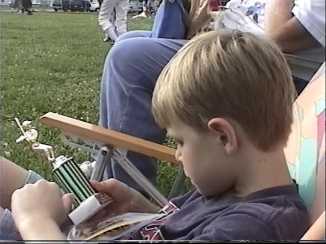 Young boy sits in a lawn chair examining a small trophy while watching a youth soccer game on a grassy field