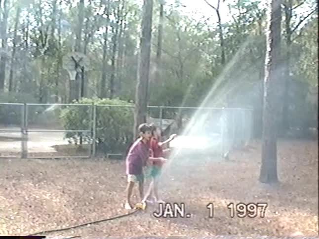 Two young boys have fun playing with a sprinkler and a water hose in their backyard in this vintage 1997 footage