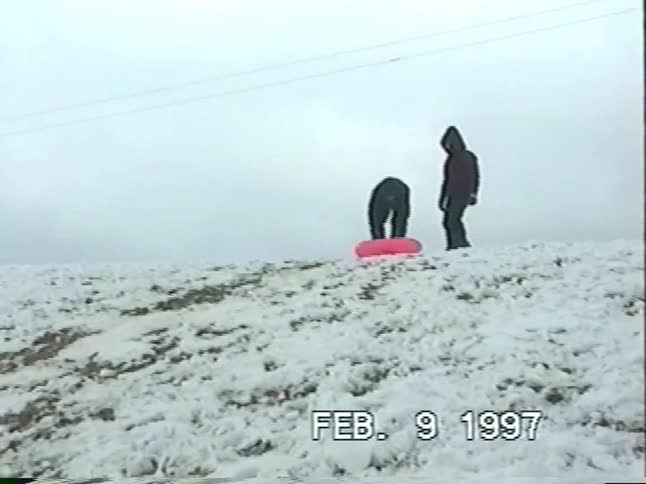 A person gets a push on a pink snow tube and slides down a snowy hill in vintage 90s footage