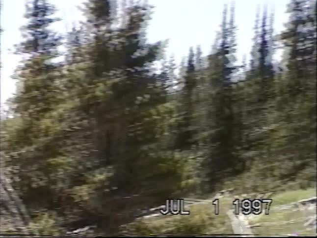 Vintage 1997 home video footage shows a group of people hiking up a grassy mountainside on a sunny day