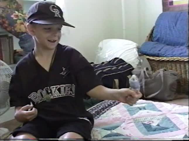 A young boy wearing a baseball cap smiles as he holds his pet parakeet on his finger in his bedroom