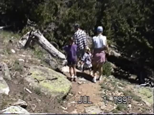 A family with two children hikes along a sunny mountain trail in vintage VHS footage from 1997