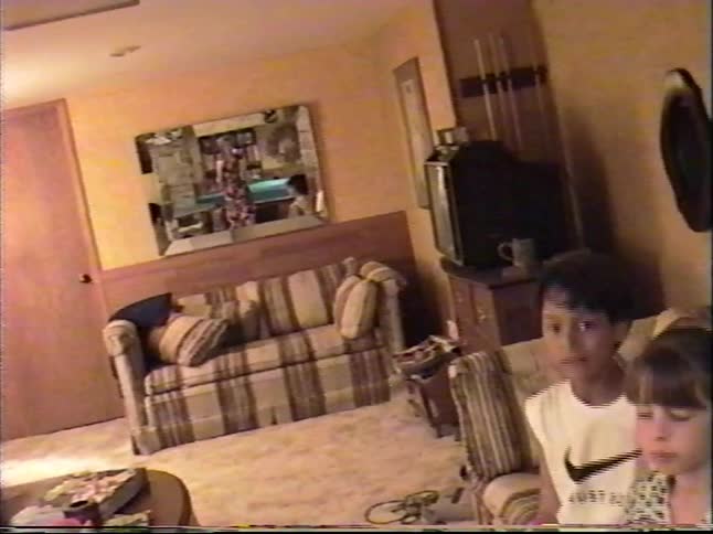 Two children wave and interact with a home video camera in a cozy living room