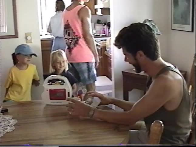A family gathers around a dining room table as one man hands another a package in this vintage home video