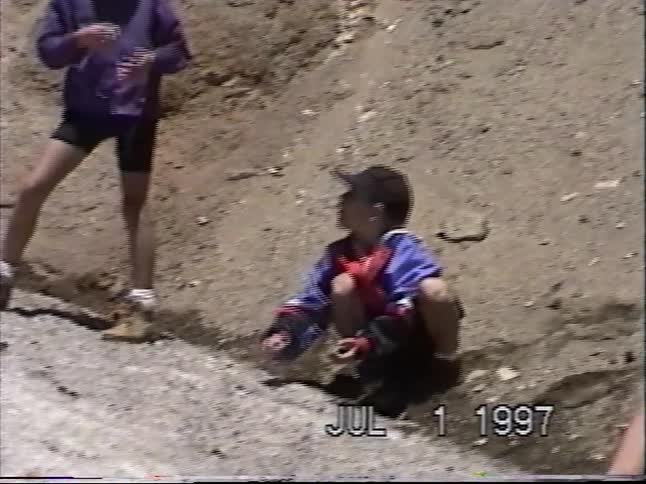 A group of kids hang out and play in a large sandy pit on a sunny day in this vintage 1990s home movie