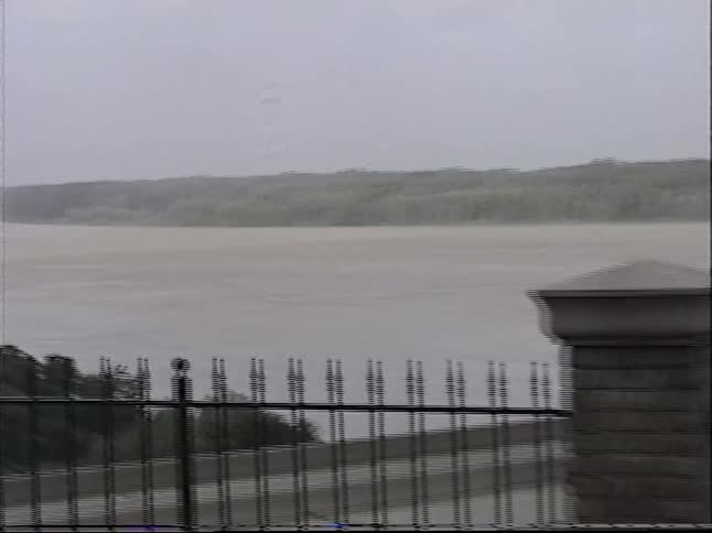 Vintage home video footage shows a man and two boys at a scenic overlook viewing two large truss bridges crossing a wide river on a cloudy day