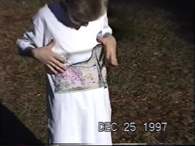 Vintage VHS footage shows a young boy happily modeling his new oversized dinosaur t-shirt on Christmas Day in 1997