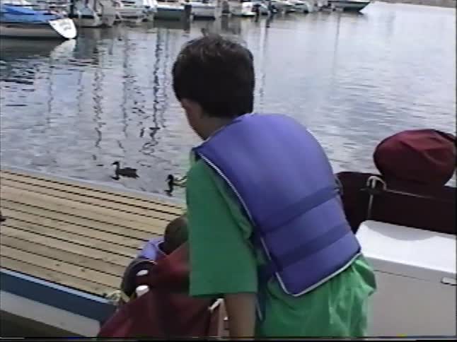 Vintage home movie footage of young children wearing life vests on a boat docked in a marina while watching ducks swim by