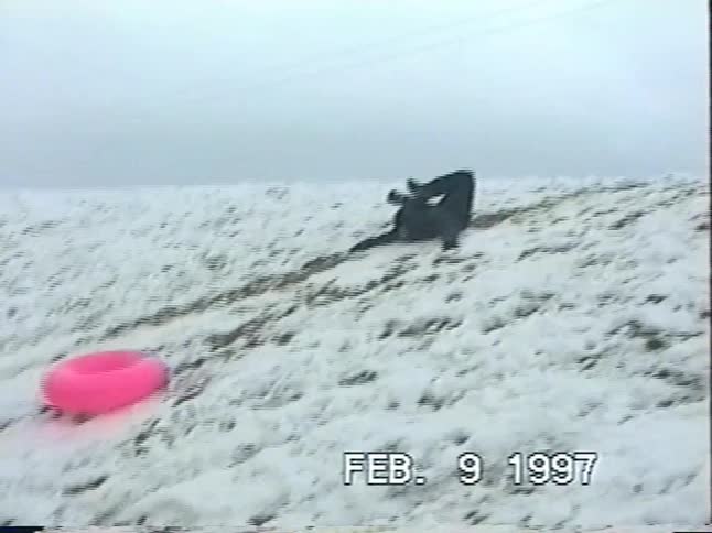 A person in a snowsuit hilariously falls before successfully sledding down a snowy hill on a pink inner tube in this vintage 1997 footage