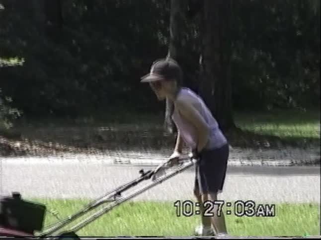 Vintage home video footage shows a young boy wearing a cap and sunglasses mowing a large lawn on a sunny day