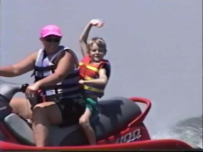 Vintage home movie footage shows a mother and her young son riding a red jet ski on a lake