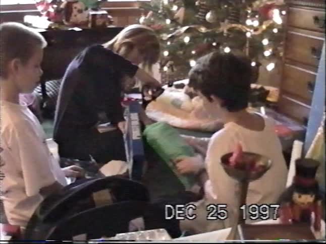 A mother and her two sons open presents together by the Christmas tree in a nostalgic home video from 1997