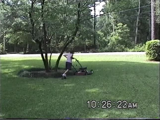 A young boy diligently mows a large green lawn with a push mower in this vintage home video footage