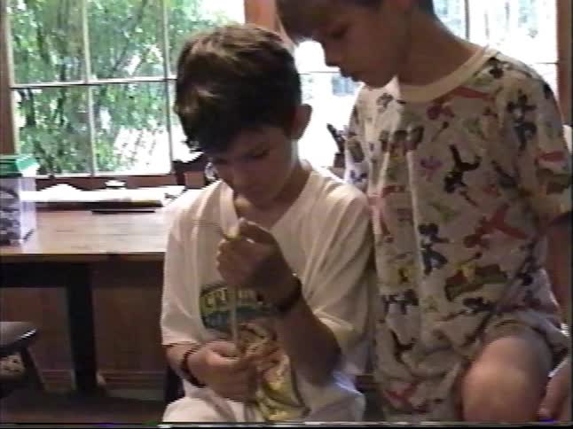 Two boys indoors gently handling a small snake near a table and aquarium in a home setting